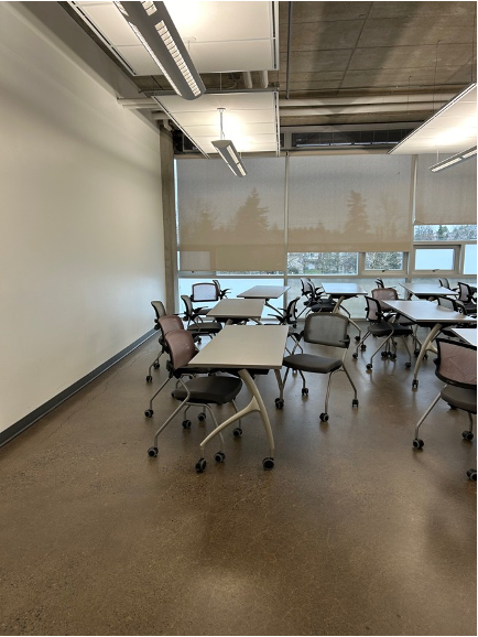 Image of Classroom 3830, large windows with blinds partially closed, large white wall, numerous long tables and chairs