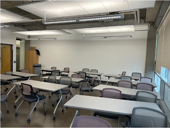 Image of Classroom 3830, large windows with blinds partially closed, large white wall, numerous long tables and chairs