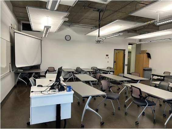 Image of Classroom 3830, large white wall, numerous long tables and chairs, screen in the left corner, instructors station