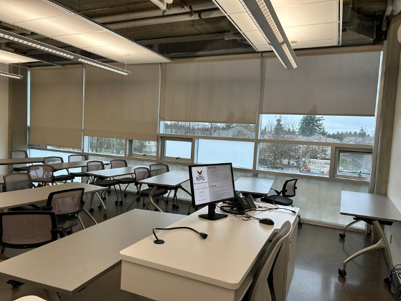 Image of Classroom 3830, wall of large windows with blinds partially down, numerous long tables and chairs, instructors station with computer