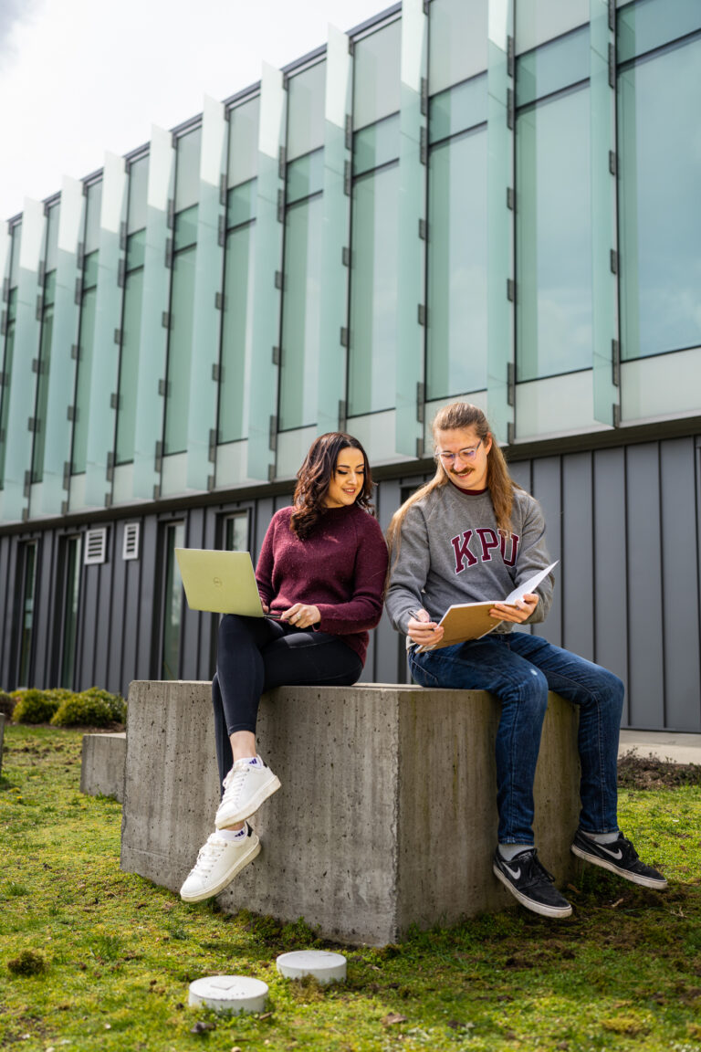 Two students sitting outside, working on laptops and sharing ideas.